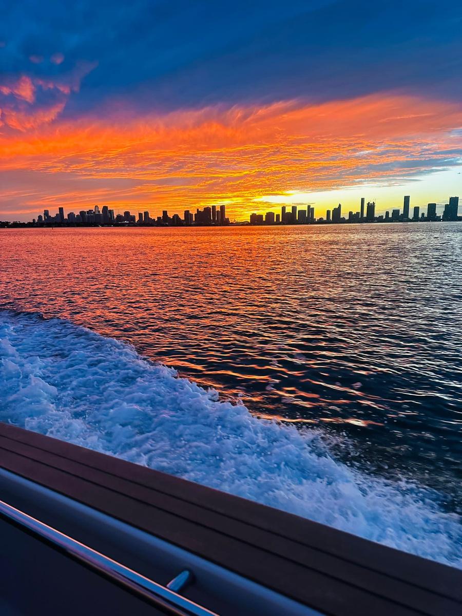 The Miami skyline in silhouette under a vivid orange and pink sunset, seen across Biscayne Bay from the teak side deck of a moving boat with its white wake churning alongside